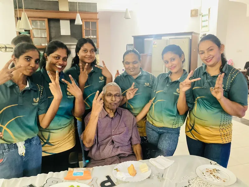 Six caregivers in team shirts pose with peace signs around a smiling resident