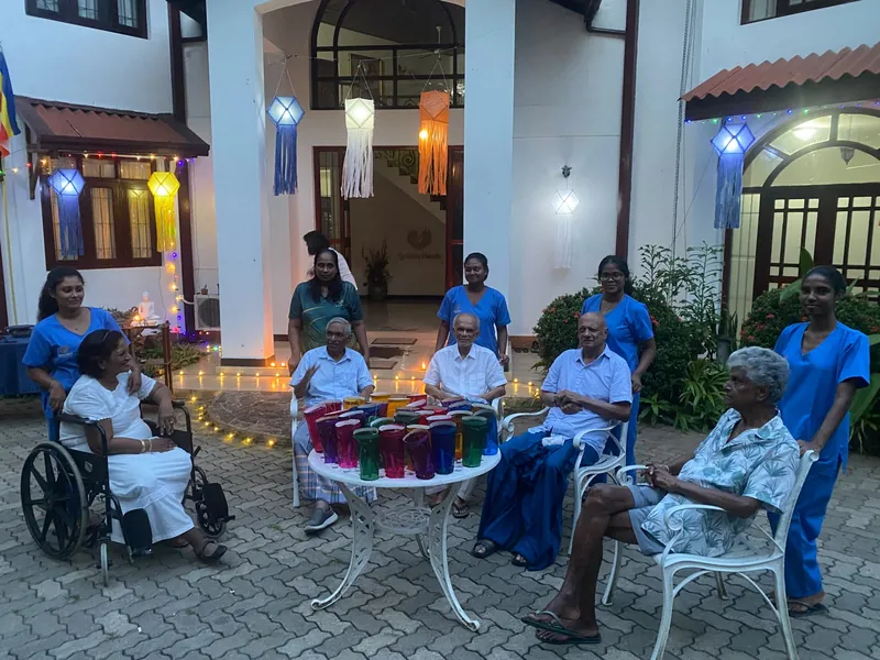 Residents and caregivers gathered around a table of lanterns in the lamplit courtyard at dusk